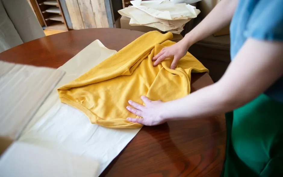 Des mains plient un t-shirt jaune sur une table en bois, une tâche du quotidien à la maison